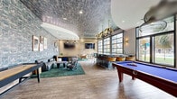 Located at 1920 Exchange Dr in Greenville, NC, this single-floor amenity lounge is a thoughtfully designed social space that seamlessly blends bold aesthetics with functional leisure areas. The interior is defined by striking textured wall coverings with an abstract, marble-like pattern in cool blue and grey tones, paired with a matching decorative ceiling treatment that adds dramatic visual depth throughout.

The space is anchored by a stunning central bar featuring an artistic, patterned facade with bar-height seating, illuminated by sculptural gold palm-leaf pendant lights that serve as a key design statement. Full-height glass garage doors line one wall, flooding the interior with natural light and offering expansive outdoor views of lush greenery and palm trees, creating a seamless indoor-outdoor ambiance.

Multiple lounge areas are thoughtfully arranged throughout the space, furnished with grey leather sofas, eclectic accent chairs, and tropical-patterned area rugs in rich forest green. Curated wall art, potted plants, and wood slat partition accents lend a warm, resort-inspired character to the interior.

A pool table positioned near the garage doors adds a recreational focal point, rounding out the space as a well-equipped social hub. Wood-look tile flooring ties all zones together, creating a cohesive and inviting atmosphere ideal for gathering and relaxation.