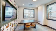 Located at 120 Matheson Ave in Charlotte, NC, this thoughtfully designed single-floor space blends modern aesthetics with vibrant, artistic character. The layout includes a meeting room, an intimate seating nook, and a bathroom, making it well-suited for collaborative or private use.

The meeting room is a standout feature, anchored by a sleek dark-topped conference table paired with stylish rattan-accented chairs. Two large black-framed windows flood the room with natural light and offer views of the surrounding streetscape. A wall-mounted display screen sits against a lively, illustrated wallpaper featuring colorful character portraits, adding a playful and creative energy to the space.

The central corridor connects the rooms with deep navy blue walls, patterned carpet tile flooring, and glass-paneled doors with dark metal frames — a clean, contemporary aesthetic that runs throughout. A cozy dining nook is tucked against a wood-paneled wall, outfitted with a green upholstered booth, a small table, and a pendant light overhead, creating a warm and intimate atmosphere.

Additional character is found in an open alcove accented with bold, graphic wallpaper, housing a single lounge chair — ideal as a quiet retreat. Recessed lighting throughout ensures a well-lit environment, while the curated artwork and décor give the space a distinctly creative and welcoming ambiance.