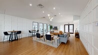 This thoughtfully designed single-floor space blends modern aesthetics with functional versatility. The centerpiece is a generous open lounge area anchored by vaulted ceilings that rise dramatically overhead, accentuating the sense of spaciousness throughout. A striking pendant light cluster with gold and globe fixtures serves as an eye-catching focal point above multiple seating arrangements, including a tan leather sofa, a gray sectional, and mid-century modern accent chairs arranged atop a large textured area rug. One wall features elegant white paneling, adding architectural texture and depth to the bright, airy interior. Warm-toned hardwood flooring flows seamlessly throughout the entire space. A wall of black-framed glass panels separates the lounge from a dedicated fitness room, which is outfitted with exercise equipment and receives abundant natural light. The kitchen area is well-appointed with crisp white shaker cabinetry, light countertops, a classic white tile backsplash, open rustic wood shelving, and a retro-style black refrigerator. A large window in the kitchen frames views of an outdoor patio area. The overall ambiance is clean, warm, and contemporary, making this space well-suited for both social gatherings and active everyday use.