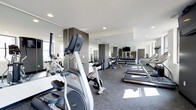 Located at 50 W Broad St in the heart of downtown Columbus, Ohio, this fitness center offers a well-equipped, single-floor workout facility designed to meet a wide range of exercise needs. The space features a clean, contemporary aesthetic with neutral tones, recessed lighting, and large windows that invite natural light throughout the room. Wall-to-wall mirrors line the space, creating an open and expansive feel while allowing users to monitor their form during workouts. The cardio section is prominently featured, housing a selection of elliptical trainers, upright cycles, and treadmills arranged to maximize floor space and user comfort. A dedicated free weights area includes a full rack of dumbbells alongside additional strength training equipment, catering to both beginner and experienced gym-goers. A wall-mounted flat-screen television adds an entertainment element to the cardio experience. The flooring consists of durable, low-pile carpet suited for high-traffic athletic use. Structural columns with dark accent panels add a modern architectural touch throughout the room. The overall ambiance is professional and energizing, making it a functional and inviting environment for daily fitness routines.