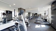 Located at 50 W Broad St in the heart of downtown Columbus, Ohio, this fitness center offers a well-equipped, single-floor workout facility designed to meet a wide range of exercise needs. The space features a clean, contemporary aesthetic with neutral tones, recessed lighting, and large windows that invite natural light throughout the room. Wall-to-wall mirrors line the space, creating an open and expansive feel while allowing users to monitor their form during workouts. The cardio section is prominently featured, housing a selection of elliptical trainers, upright cycles, and treadmills arranged to maximize floor space and user comfort. A dedicated free weights area includes a full rack of dumbbells alongside additional strength training equipment, catering to both beginner and experienced gym-goers. A wall-mounted flat-screen television adds an entertainment element to the cardio experience. The flooring consists of durable, low-pile carpet suited for high-traffic athletic use. Structural columns with dark accent panels add a modern architectural touch throughout the room. The overall ambiance is professional and energizing, making it a functional and inviting environment for daily fitness routines.