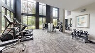 Located at 1100 Hammond Dr in Sandy Springs, GA, this fitness center offers a well-appointed, single-floor exercise space designed with both function and aesthetics in mind. The room's most striking architectural feature is its dramatic double-height ceiling, which creates an open, airy atmosphere and contributes to an energizing workout environment. Exposed concrete columns punctuate the space, adding an industrial-modern character that complements the clean white walls and warm wood accents.

Floor-to-ceiling windows line multiple walls, flooding the interior with abundant natural light and providing views of the surrounding greenery — a refreshing backdrop during any workout. The textured, patterned carpet flooring adds visual warmth while providing a practical surface underfoot.

The fitness center is comprehensively equipped to support a wide range of training goals. Cardio equipment includes elliptical trainers, stair climbers, and treadmills arranged to take full advantage of the natural light and outdoor views. The strength training area features a variety of cable and resistance machines, a power rack, adjustable benches, kettlebells, and a medicine ball station. Mirrored walls in select areas enhance the sense of space and allow users to monitor their form. The overall ambiance is modern, bright, and motivating.