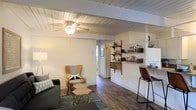 This single-floor space showcases a thoughtfully designed open-plan layout that seamlessly connects the living and dining areas. The ceiling is a standout feature, finished with white-painted wood planks and exposed beams that lend a rustic yet clean aesthetic. A ceiling fan is centrally positioned, enhancing both function and style.

The living area is cozy and well-defined, accommodating a sofa and an accent armchair with a coordinating ottoman arranged over a patterned area rug. Rich, dark hardwood flooring runs throughout the space, grounding the design with warmth and continuity.

Adjacent to the living area, a compact bar or dining counter with a leather-upholstered stool provides a casual space for dining or work. Open wooden shelving units mounted along the wall serve as both storage and display, holding glassware, small appliances, and decorative items — effectively defining a kitchenette zone without the need for full cabinetry.

The neutral white walls keep the space feeling bright and airy, while the mix of wood tones and modern furnishings creates a balanced, inviting ambiance. A hallway visible in the background suggests additional rooms beyond the main living area.
