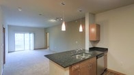 This single-floor space is designed with an open-concept layout that promotes a seamless flow between the kitchen and living areas. The kitchen serves as the centerpiece, featuring a central island topped with dark granite countertops and a double stainless steel sink. Warm wood cabinetry provides ample storage, while two elegant pendant lights hang above the island, adding both style and focused task lighting. Upper cabinets along the wall extend the kitchen's storage capacity, complemented by additional dark granite counter space. A track lighting fixture overhead further enhances the kitchen's illumination. The open layout transitions naturally into a spacious carpeted living area, which benefits from natural light streaming in through a large window. Neutral wall tones throughout create a calm, cohesive ambiance, while the combination of recessed lighting and pendant fixtures ensures the space is well-lit at all times. A hallway visible in the background hints at additional rooms beyond, adding to the overall functionality of the floor plan. The thoughtful design balances practicality with contemporary aesthetics, making this a comfortable and visually appealing space.