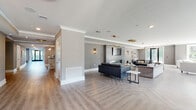 This elegantly designed single-floor amenity space offers an expansive, open-concept layout that seamlessly blends comfort and contemporary style. The flooring throughout is a standout feature — light-toned hardwood laid in a classic herringbone pattern that adds visual depth and warmth to the entire space.

The main lounge area is generously proportioned, divided into multiple seating vignettes furnished with a curated mix of leather sofas, tufted settees, velvet accent chairs in navy and cream, and sleek marble-top coffee tables. A focal point of the lounge is a floor-to-ceiling shiplap accent wall in muted blue-grey, housing a linear gas fireplace and a mounted flat-screen television, flanked by elegant cylindrical wall sconces. Gold-tone circular pendant lights hang from the ceiling, adding a warm, upscale ambiance throughout.

Large floor-to-ceiling windows line multiple walls, flooding the interior with natural light and offering views of the surrounding greenery. A dedicated dining and flex seating area sits adjacent, ideal for casual gatherings or individual use.

Near the entrance, a built-in wet bar with dark cabinetry and a marble-look tile backsplash provides a convenient refreshment station. Wide hallways connect to additional corridors and adjacent spaces, all finished with crown molding and consistent neutral grey tones that unify the design throughout.