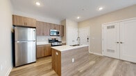 This single-floor unit features a thoughtfully designed open-concept layout that seamlessly connects the kitchen and living areas. The kitchen is a functional centerpiece, anchored by a central island with a double undermount sink, pull-down faucet, and light quartz countertops. Warm-toned wood cabinetry with brushed metal hardware lines the walls, complemented by a full suite of stainless steel appliances including a gas range, over-the-range microwave, refrigerator, and dishwasher. Light wood-style luxury vinyl plank flooring extends throughout the kitchen and living areas, adding warmth and continuity.

The spacious living area benefits from multiple windows and a sliding glass door dressed with vertical blinds, allowing ample natural light to fill the space. Neutral beige walls throughout create a clean, versatile backdrop.

The bedroom features soft carpeting, dual windows with street views, and a walk-in closet with built-in shelving. A full bathroom is finished with large-format tile flooring in a neutral tone and includes a soaking tub with shower surround. Recessed lighting throughout the unit completes the modern, move-in-ready aesthetic.