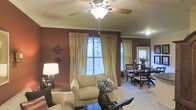 This single-floor space offers an inviting, open-concept layout that seamlessly connects the living and dining areas. The living room is anchored by plush, neutral-toned seating and a dark wood coffee table, creating a comfortable and functional focal point. Rich, warm brown walls add depth and coziness to the space, complemented by elegant crown molding that frames the ceiling throughout. A ceiling fan with a light fixture provides both comfort and ambient illumination.

Natural light filters in through multiple windows dressed with flowing, gold-toned curtains over horizontal blinds, softening the overall atmosphere. Decorative wall accents, including ornate metal pieces, add character to the living area. A table lamp provides additional warm lighting in the corner.

The adjacent dining area is visible from the living room, furnished with a dark wood table and matching chairs, making it ideal for everyday dining. A decorative floral arrangement serves as a centerpiece, adding a touch of elegance. The continuous carpeted flooring unifies both areas, contributing to the space's warm and cohesive aesthetic.