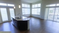 This single-floor space showcases a thoughtfully designed open-concept layout that connects the kitchen and living areas into one cohesive environment. The kitchen features a curved island with a light-toned stone countertop, an undermount double sink with a modern gooseneck faucet, and dark espresso cabinetry that provides a striking contrast against the lighter surroundings. Pendant lighting hangs directly above the island, adding focused illumination and a contemporary aesthetic touch.

The living area extends naturally from the kitchen, finished with warm wood-tone flooring that runs throughout the space, contributing to a clean and unified look. Large windows along two walls flood the interior with abundant natural light, enhancing the open and airy feel of the room. The windows are fitted with horizontal blinds, offering flexible light control. Neutral gray-white walls keep the space feeling bright and versatile. The overall ambiance is modern and welcoming, with clean lines, quality finishes, and an efficient flow that makes the space feel both functional and comfortable.