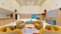 Located at 7510 N Oak Trafficway in Gladstone, Missouri, this single-floor space serves as a welcoming lobby and reception area designed with a refined, contemporary aesthetic. The standout architectural feature is the dramatically high coffered ceiling, which lends an open, airy quality and is adorned with a sculptural multi-bulb chandelier that serves as a central focal point. Warm natural oak wood paneling lines the walls, creating a cohesive and inviting atmosphere throughout.

The main lounge area is anchored by a large, curved white sectional sofa paired with a sculptural wood coffee table, complemented by plush, rounded amber-toned armchairs arranged around layered area rugs. This seating arrangement is designed to encourage comfortable gathering and conversation. Terrazzo tile flooring extends across the space, adding texture and a sense of continuity.

An adjacent reception area features a built-in wooden desk with clean, modern lines, a beadboard ceiling detail, and ample counter space. Large windows and glass entry doors allow generous natural light to fill the space and provide clear views of the surrounding outdoor environment. A secondary lounge nook near the entrance offers additional seating with benches and accent chairs. The overall ambiance is warm, modern, and functional, balancing professional utility with welcoming design.