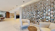 The Forte's lobby in Hackensack, NJ, makes an immediate impression with its expansive, open layout spanning a substantial footprint across a single floor. The entrance features floor-to-ceiling glass doors and large windows that flood the interior with natural light while offering views of the surrounding streetscape, creating a welcoming and airy atmosphere from the moment you step inside.

The design is anchored by large-format white porcelain tile flooring that runs seamlessly throughout, complemented by crisp white ceilings with sleek recessed and surface-mounted lighting. A dramatic textured metallic accent wall serves as a striking focal point in the lounge area, where contemporary seating arrangements with neutral-toned chairs and a warm wood coffee table invite residents to relax.

A refined conference or co-working table — crafted from rich walnut and surrounded by cognac leather and brass-framed chairs — sits beneath elegant globe pendant lights, bathed in natural light from oversized windows. Warm wood slat paneling lines the elevator vestibule, framing stainless steel elevator doors with polished sophistication.

The lobby's reception desk is clad in veined marble stone, bearing The Forte's branded signage, and is flanked by warm wood paneling and a dedicated package room. A well-organized mailbox station with marble surround and under-lighting completes the thoughtfully appointed space, delivering a seamless blend of style and practicality.