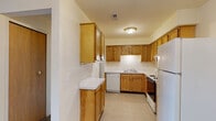 This single-floor unit is thoughtfully laid out across several interconnected rooms, offering a practical and comfortable living environment. The kitchen serves as a functional centerpiece, featuring warm honey-toned wood cabinetry extending to the upper walls for ample storage, white appliances including a full-size refrigerator, dishwasher, and range, and light-toned vinyl flooring. A peninsula counter provides additional workspace, while a pass-through window connects the kitchen to the carpeted living area, creating an open, communicative flow between spaces. A geometric gold chandelier adds a modern decorative touch above the living and dining areas. The living room is generously sized, carpeted in neutral tones, and benefits from natural light through a large window dressed with vertical blinds. The bathroom is clean and well-appointed, with tile flooring, a tub and shower combination with white tile surround, a vanity with wood cabinetry, and a large mirror with overhead lighting. Adjacent to the bathroom, a spacious walk-in closet provides built-in shelving, hanging rods, and additional storage compartments, offering excellent organization potential throughout the unit.