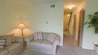 This two-floor space presents a straightforward, functional layout with a calm, neutral aesthetic throughout. The main level features a carpeted living area adorned in soft, muted tones, providing a comfortable and understated atmosphere. A plush loveseat with accent pillows anchors the sitting area, which is complemented by light-colored walls that enhance the sense of openness. Overhead, a ceiling-mounted light fixture provides ambient illumination, while a table lamp adds a warm supplementary glow. A central wall partially divides the living space from the adjoining hallway, which leads toward the entryway and the staircase accessing the upper floor. The staircase, visible from the main living area, features dark-toned treads that contrast with the lighter surroundings. The entryway at the end of the hall includes a door with an adjacent sidelight, allowing natural light to filter into the corridor. The carpeting extends throughout the main level, contributing to a cohesive and quiet interior feel. Ceiling vents indicate a central HVAC system, ensuring climate control across both floors. Overall, the space offers a clean, practical environment with a neutral palette.