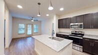 Located at 329–369 E Mars Rd in Wilmer, Texas, this single-story unit offers a contemporary design with a cohesive neutral color palette and light wood-style flooring throughout. The open-concept layout connects the kitchen and living area, creating a functional and visually spacious environment.

The kitchen is a standout feature, centered around a large island with a granite countertop and an undermount sink, illuminated by pendant lighting overhead. Dark espresso cabinetry with sleek hardware lines the walls, providing ample storage, while stainless steel appliances—including a range, microwave, and refrigerator—complete the modern aesthetic. Recessed lighting adds consistent illumination across the space.

The living area accommodates comfortable furnishing arrangements and is brightened by natural light from a set of double doors and adjacent windows, with a ceiling fan overhead. The bedroom includes a walk-in closet with built-in shelving for organized storage.

The bathroom is well-appointed with a double-sink vanity featuring granite countertops, dark cabinetry, a tall storage tower, and large mirrors framed by vanity lighting. A tub-shower combination with tiled surround and built-in shelving completes the bath.