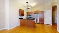 This single-floor unit is defined by warm, natural hardwood flooring that runs consistently throughout the entire space, creating a cohesive and inviting atmosphere. The kitchen serves as a central highlight, designed with rich medium-toned wood cabinetry that extends to the upper walls, providing generous storage. Dark granite countertops offer a striking contrast against the cabinetry, while a peninsula with matching countertop and beadboard detailing adds functional workspace and a natural division between the kitchen and living areas. A stylish multi-light pendant chandelier anchors the kitchen visually. Stainless steel appliances, including a refrigerator, electric range, and built-in microwave, deliver a modern, polished finish. The living areas are bright and open, featuring neutral-toned walls that complement the warm flooring beneath. Bedroom spaces are well-proportioned, each benefiting from large windows fitted with horizontal blinds that allow natural light to filter in while offering privacy. Clean white baseboards frame each room neatly. The overall layout, as reflected in the floor plan, flows logically from the kitchen through the living and bedroom spaces, making efficient use of the available area.