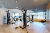 Located at 4845 Ch De La Côte-Saint-Luc in Montréal, this fitness facility offers a thoughtfully designed single-floor exercise room that provides a comprehensive workout environment. The space is defined by its curved glass window wall on one side, flooding the room with natural light and offering open views of the surrounding exterior — creating an airy, motivating atmosphere for exercise.

The light hardwood flooring throughout contributes to a clean, modern aesthetic, while recessed ceiling lighting ensures the space is well-lit at all times. The ceiling features a dual-tone design, with portions finished in dark grey that add a contemporary, dynamic character to the room.

The exercise room is equipped with a diverse selection of fitness equipment to support both strength training and cardiovascular workouts. Strength training options include a multi-function cable machine, a weight bench with attachments, and a rack of free-weight dumbbells. Cardio equipment includes treadmills, an elliptical trainer, a stationary bike, and a rowing machine. Stability balls and yoga mats are also available for flexibility and core training. A full-length mirror on one wall enhances the sense of space and allows users to monitor their form. A dedicated storage unit and posted gym regulations round out the practical amenities of this well-organized facility.