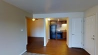 This single-floor space features an open, flowing layout unified by rich dark hardwood flooring throughout. The main living area connects seamlessly to adjoining rooms, offering a sense of continuous, functional space. A ceiling fan is installed in one of the connecting rooms, providing both airflow and a practical design element.

The kitchen is nestled within a recessed nook, defined by dark wood cabinetry that extends to the upper walls, providing ample storage. Black appliances, including a full-sized refrigerator and dishwasher, complement the cabinetry for a cohesive, contemporary look. A under-cabinet light illuminates the countertop workspace, which features a sink positioned along the back wall. Recessed lighting adds additional brightness to the kitchen area.

The layout benefits from an open passage between the living and kitchen spaces, creating an interconnected feel ideal for everyday living. White walls and trim contrast the darker flooring and cabinetry, keeping the overall aesthetic balanced and clean. A white door on the right side of the main area provides access to an additional room or exterior. The space presents a straightforward, well-organized interior suited for comfortable, practical living.