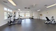 This fitness center is a spacious, single-floor facility designed to accommodate a comprehensive workout experience. The room is defined by crisp white walls adorned with crown molding, and a durable dark rubberized flooring that spans the entire space, providing both safety and comfort during exercise. Ceiling fans and recessed fluorescent lighting ensure the environment remains well-lit and well-ventilated throughout.

Natural light streams in through multiple sets of windows along the walls, offering views of the surrounding greenery and creating an open, inviting atmosphere. French doors on one side provide an additional entry point and further enhance the sense of airiness within the space.

The facility is outfitted with a variety of cardio equipment, including treadmills, elliptical trainers, and stationary bikes, thoughtfully arranged to allow for easy movement between machines. A dedicated free-weight area features a dumbbell rack and a weight bench, supporting strength training routines. Wall-mounted televisions are positioned at multiple points throughout the room, allowing users to stay entertained during their workouts.

A water cooler and informational bulletin board are conveniently placed near the entrance, and the generous open floor plan leaves ample room for stretching or floor-based exercises. The overall design is clean, functional, and welcoming.