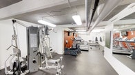 This fitness room offers a practical and well-organized space for exercise, designed to accommodate a range of workout needs. The room features an industrial aesthetic, with exposed ductwork and concrete ceilings overhead, complemented by bright fluorescent lighting that ensures the space is well-lit throughout. The flooring consists of durable dark rubber matting, ideal for high-traffic gym use and providing a stable surface for equipment and exercise.

Along one side of the room, a multi-station cable and weight machine offers a wide range of strength training options. A row of cardio equipment — including treadmills, an elliptical, and a stationary bike — lines the center and far wall, providing ample options for aerobic workouts. An accent wall painted in a warm terracotta tone adds a subtle visual element to the otherwise neutral color palette. Toward the back of the room, a mirrored wall and bench seating area provide space for stretching or free weight exercises. The layout is efficient and straightforward, making the most of the available space.