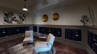 Located at 20 Romney St in Charleston, SC, this thoughtfully designed mail room blends practical functionality with refined aesthetic details. The space is divided into two distinct zones connected by a short staircase, each finished with its own flooring — classic penny-tile in one area and rich hardwood in the other — adding visual interest and character throughout.

Banks of matte black individual mailboxes line the walls on multiple sides, providing ample mail storage for residents. A substantial dark-topped table with ornate industrial-style base occupies the center of the tiled zone, serving as a convenient surface for sorting mail and packages. Overhead, pendant lights with vintage-inspired shades hang from the ceiling alongside exposed wooden beams, casting warm, focused illumination throughout the room.

The hardwood-floored section offers a more lounge-like atmosphere, featuring two upholstered mid-century-style armchairs arranged on a patterned area rug — an inviting spot to pause. Decorative oversized brass disc wall art adds a bold, sculptural element above the mailboxes, complementing the space's overall sophisticated tone. Artwork and potted plants are positioned throughout, softening the utilitarian nature of the room. A wall-mounted television and mirror further enhance the space's amenity-rich character.