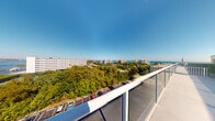 This single-floor property is defined by its remarkable outdoor terrace, which serves as a standout architectural feature. The terrace is generously proportioned, wrapping around the exterior and finished with clean, light-toned pavers that reflect the abundant Florida sunshine. A sleek glass and metal railing system runs the length of the terrace, ensuring unobstructed sightlines while maintaining a modern, refined aesthetic.

From the terrace, the views are a prominent highlight. Lush, mature tree canopies stretch across the foreground, giving way to neighboring low-rise buildings and, beyond them, a clear view of the ocean horizon under an expansive blue sky. The coastal setting is immediately apparent, with the Atlantic visible in the distance and the surrounding landscape characterized by tropical greenery typical of South Florida.

The terrace connects to the interior through glass-paneled doors, suggesting a seamless indoor-outdoor relationship designed to take full advantage of the coastal environment. The overall ambiance is bright, open, and airy, with the elevation of the space providing a sense of privacy and commanding presence above the surrounding neighborhood. The combination of the panoramic views, modern railing design, and spacious outdoor area makes this terrace a central and defining element of the property.