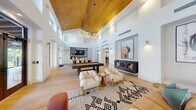 This expansive single-floor amenity space is defined by its dramatic architectural presence, centered around a soaring vaulted ceiling clad in warm-toned wood planking. The ceiling's height is further emphasized by a clerestory window band that wraps the upper perimeter, flooding the interior with abundant natural light and creating an airy, open atmosphere throughout.

Light-washed hardwood flooring extends across the entire space, unifying its distinct functional zones. The main lounge area features a curated arrangement of cream upholstered armchairs with natural wood frames, a leather sofa, and patterned area rugs, all set against crisp white walls adorned with bold, oversized framed artwork displayed in recessed niches with dedicated sconce lighting.

A dedicated game zone accommodates both a full-size billiards table and a shuffleboard table, positioned to take full advantage of the room's generous proportions. Large steel-framed windows and glass doors open directly to an outdoor terrace, offering views of lush landscaping and a pool area beyond.

A secondary area includes a kitchen-adjacent nook finished with striking scalloped mosaic tile, built-in white cabinetry with matte black hardware, and a floating shelf—ideal as a serving station. The overall ambiance is modern, warm, and resort-inspired.