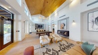 This expansive single-floor amenity space is defined by its dramatic architectural presence, centered around a soaring vaulted ceiling clad in warm-toned wood planking. The ceiling's height is further emphasized by a clerestory window band that wraps the upper perimeter, flooding the interior with abundant natural light and creating an airy, open atmosphere throughout.

Light-washed hardwood flooring extends across the entire space, unifying its distinct functional zones. The main lounge area features a curated arrangement of cream upholstered armchairs with natural wood frames, a leather sofa, and patterned area rugs, all set against crisp white walls adorned with bold, oversized framed artwork displayed in recessed niches with dedicated sconce lighting.

A dedicated game zone accommodates both a full-size billiards table and a shuffleboard table, positioned to take full advantage of the room's generous proportions. Large steel-framed windows and glass doors open directly to an outdoor terrace, offering views of lush landscaping and a pool area beyond.

A secondary area includes a kitchen-adjacent nook finished with striking scalloped mosaic tile, built-in white cabinetry with matte black hardware, and a floating shelf—ideal as a serving station. The overall ambiance is modern, warm, and resort-inspired.