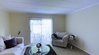 This two-floor residence offers a comfortable and functional living environment with notable design details throughout. The living room serves as a central gathering space, featuring wall-to-wall carpeting that adds warmth underfoot. A large sliding glass door dominates one wall, fitted with vertical blinds that allow for adjustable natural light control. The door provides direct access to an outdoor patio area, where outdoor seating is visible, creating a seamless indoor-outdoor connection. The living room accommodates generous seating, including a cushioned armchair, and is styled with a round glass coffee table atop a dark area rug, adding visual contrast to the light-toned carpet. A glass-top side table with a metal frame provides additional surface space. The walls are finished in neutral tones, complementing the overall bright and clean aesthetic. The ceiling features subtle architectural detailing with a linear panel design, adding a modest structural element to the space. Crown molding along the upper walls contributes a polished finish. The overall ambiance of this home is light, open, and welcoming, with practical features suited for everyday living across both floors.