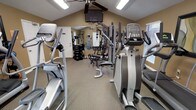 This fitness center offers a comprehensive workout environment designed to accommodate a wide range of exercise needs. The space features vaulted ceilings that lend an open, airy feel, complemented by a ceiling fan that aids in air circulation during workouts. Natural light filters in through windows fitted with blinds, adding brightness to the warm, neutral-toned walls.

The cardio section is well-appointed with multiple elliptical trainers and a treadmill, providing ample options for aerobic exercise. A ceiling-mounted flat-screen television is positioned for easy viewing from the cardio equipment, enhancing the workout experience. Toward the back of the space, a multi-station strength training machine serves as the centerpiece of the weight training area, surrounded by a rack of free weights and additional resistance equipment. The flooring throughout is a durable, low-pile carpet suited for fitness use.

A standard entry door provides access to the space, and the overall layout is thoughtfully arranged to allow comfortable movement between equipment. The combination of cardio machines, free weights, and multi-use strength equipment makes this a versatile and fully functional fitness facility.