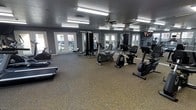 This fitness center offers a thoughtfully designed, open workout space that accommodates a wide range of exercise routines. The room is finished with durable carpet tile flooring and features a textured ceiling with flush-mount fluorescent lighting, ensuring bright, even illumination throughout. A ceiling fan adds air circulation, enhancing comfort during workouts. The walls are painted in a cool grey tone, creating a focused and energizing atmosphere. Large French-style doors and windows line the walls, inviting generous natural light and providing views of the surrounding greenery, helping to create an open and airy feel. Full-length mirrors are strategically positioned to expand the sense of space and allow users to monitor their form. The facility is equipped with a comprehensive selection of exercise equipment, including treadmills, upright and recumbent stationary bikes, a cable resistance machine, and additional strength and cardio units. A wall-mounted television is also present, providing entertainment during workouts. The overall layout is open and well-organized, allowing ample room to move freely between stations, making this fitness center a fully functional and welcoming environment for residents or members looking to maintain an active lifestyle.