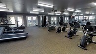 This fitness center offers a thoughtfully designed, open workout space that accommodates a wide range of exercise routines. The room is finished with durable carpet tile flooring and features a textured ceiling with flush-mount fluorescent lighting, ensuring bright, even illumination throughout. A ceiling fan adds air circulation, enhancing comfort during workouts. The walls are painted in a cool grey tone, creating a focused and energizing atmosphere. Large French-style doors and windows line the walls, inviting generous natural light and providing views of the surrounding greenery, helping to create an open and airy feel. Full-length mirrors are strategically positioned to expand the sense of space and allow users to monitor their form. The facility is equipped with a comprehensive selection of exercise equipment, including treadmills, upright and recumbent stationary bikes, a cable resistance machine, and additional strength and cardio units. A wall-mounted television is also present, providing entertainment during workouts. The overall layout is open and well-organized, allowing ample room to move freely between stations, making this fitness center a fully functional and welcoming environment for residents or members looking to maintain an active lifestyle.