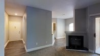 This single-floor residence offers a thoughtfully designed open layout that promotes a sense of spaciousness and easy flow between living areas. The main living space is anchored by a sleek fireplace with a contrasting surround, creating a natural focal point for the room. Soft, neutral-toned carpet underfoot adds warmth and comfort to the living area, while hardwood-style flooring transitions into the adjacent spaces, providing both durability and visual continuity. The walls are painted in a calming gray tone, complemented by crisp white baseboards that give the interior a clean, modern finish. Natural light filters through a window in the adjoining area, brightening the space and enhancing its open feel. A partial view into what appears to be a kitchen or utility area reveals built-in shelving and countertop space, hinting at practical storage and functionality. A hallway extends toward additional rooms, suggesting a well-organized floor plan. Recessed ceiling vents indicate a central HVAC system, contributing to year-round comfort. Overall, the space presents a move-in-ready, contemporary aesthetic well-suited for everyday living.