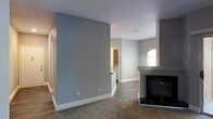 This single-floor residence offers a thoughtfully designed open layout that promotes a sense of spaciousness and easy flow between living areas. The main living space is anchored by a sleek fireplace with a contrasting surround, creating a natural focal point for the room. Soft, neutral-toned carpet underfoot adds warmth and comfort to the living area, while hardwood-style flooring transitions into the adjacent spaces, providing both durability and visual continuity. The walls are painted in a calming gray tone, complemented by crisp white baseboards that give the interior a clean, modern finish. Natural light filters through a window in the adjoining area, brightening the space and enhancing its open feel. A partial view into what appears to be a kitchen or utility area reveals built-in shelving and countertop space, hinting at practical storage and functionality. A hallway extends toward additional rooms, suggesting a well-organized floor plan. Recessed ceiling vents indicate a central HVAC system, contributing to year-round comfort. Overall, the space presents a move-in-ready, contemporary aesthetic well-suited for everyday living.