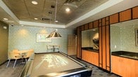 This single-floor game room is thoughtfully designed as a dedicated entertainment space. At its center sits a full-sized pool table, illuminated by a classic pendant lamp that draws the eye and anchors the room. The ceiling features ornate decorative tiles with a geometric pattern, complemented by recessed lighting that provides warm, even illumination throughout.

The walls are finished with a textured, stone-like surface that adds depth and visual interest to the space. A standout architectural element is the built-in wood cabinetry unit along one wall, framed in a grid-style design with warm toned wood panels and a dark countertop, offering both storage and display space. A decorative sculptural piece rests atop the unit, adding an artistic touch.

In the background, a casual dining nook with a small table and chairs provides a comfortable spot for guests to gather. A framed piece of artwork adorns the nearby wall. The flooring features a patterned area rug that ties the design elements together, creating a cohesive, inviting atmosphere ideal for socializing and recreation.