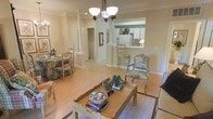 This single-floor residence offers a welcoming and cohesive open-concept layout that seamlessly connects the main living areas. The living room serves as the heart of the home, anchored by a classic coffee table, an elegant upholstered armchair with ornate wooden detailing, and a comfortable sofa, all resting on warm hardwood-style flooring that runs throughout the space. A decorative chandelier with frosted glass shades hangs overhead, casting a soft, inviting glow across the room. A decorative column adds an architectural accent, subtly dividing the living and dining areas while maintaining an open, airy feel. The dining area accommodates a charming table and chair set, ideal for casual meals or entertaining. Crown molding lines the ceilings, adding a refined touch to the overall aesthetic. The kitchen features a convenient pass-through bar counter with barstool seating, white cabinetry, and white appliances, offering both functionality and easy interaction with the adjoining living spaces. Neutral, warm-toned walls throughout the home contribute to a calm and comfortable ambiance, while the thoughtful layout maximizes flow and usability across every area of this well-appointed single-level residence.