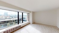 This single-floor unit offers a clean, neutral aesthetic throughout, with each space thoughtfully laid out for comfortable everyday living. The main living area is a standout feature, characterized by plush wall-to-wall carpeting and a large bank of floor-to-ceiling windows with black metal frames. These expansive windows flood the room with natural light and frame sweeping views of the surrounding cityscape, including neighboring rooftops, architectural landmarks, and a distant skyline — creating a bright and open atmosphere.

The galley-style kitchen is efficiently designed, with white cabinetry lining both sides, light countertops, and essential appliances including a full-size refrigerator and an electric range with overhead ventilation. Durable vinyl flooring and ceiling-mounted lighting give the kitchen a practical, clean finish.

The bathroom is well-appointed with a full-size soaking tub and shower combination, accented by neutral tile surround. A wide vanity mirror spans the length of the sink area, complemented by overhead strip lighting for a bright and functional space.

A dedicated closet with a full-width hanging rod provides ample storage. Throughout the unit, the consistent use of soft whites and warm neutrals creates a calm, cohesive ambiance well-suited for a variety of lifestyles.