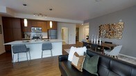 This single-floor residence offers a thoughtfully designed open-concept layout that seamlessly connects the kitchen, dining, and living areas into one cohesive and functional space. The kitchen is a standout feature, showcasing warm wood cabinetry, stainless steel appliances including a built-in microwave and refrigerator, and a sleek white countertop island with bar seating — ideal for casual dining or entertaining. Track lighting and a pendant light above the island provide focused illumination throughout the kitchen. The living area is anchored by a bold black leather sofa accented with layered throw pillows, lending a modern yet inviting atmosphere. Warm-toned hardwood flooring runs throughout the main living space, adding continuity and warmth. The dining area accommodates a full-sized table with upholstered chairs, complemented by decorative accents including sculptural candleholders and a geometric wall piece that adds visual interest. Hallways lead to additional private rooms, offering a natural separation between the social and private areas of the home. The overall design balances contemporary finishes with functional living, creating a polished and comfortable environment.