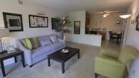 This single-story space offers an open-concept layout that seamlessly connects the living area to the kitchen. The living room is carpeted and comfortably sized, providing ample room for seating arrangements centered around a dark wood coffee table. A lavender sofa and a chartreuse accent chair anchor the space, complemented by colorful framed artwork on the neutral-toned walls and a tall indoor palm that adds a natural, organic touch. A ceiling fan overhead ensures comfortable air circulation throughout the open area. The half-wall partition elegantly separates the living space from the kitchen, which features warm wood cabinetry and a functional countertop workspace visible from the main living area. The kitchen's pass-through design enhances the sense of openness and connectivity between spaces. Decorative wall accents and thoughtfully placed accessories contribute to the cohesive, welcoming ambiance. The consistent neutral wall palette and carpeted flooring throughout create a unified aesthetic that ties the entire space together.