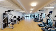 The fitness center at 360 Huguenot St in New Rochelle, NY is a thoughtfully designed, single-level facility offering a comprehensive workout environment. The space is organized into distinct functional zones, each featuring light-toned hardwood-style flooring or rubberized gray flooring suited to specific training activities.

The cardio area is generously equipped with treadmills, ellipticals, stationary bikes, stair climbers, and rowing machines, including ROW Aktiv rowers. This section benefits from natural light via multiple windows and is illuminated by a combination of flush drum pendant fixtures and linear fluorescent lighting, contributing to a clean, bright atmosphere.

The strength and functional training zones include selectorized resistance machines, a multi-functional cable system, a power rack with a flat bench, and a dumbbell rack. A dedicated functional training area features suspension trainers, heavy bags, kettlebells, and battle ropes arranged around a central rig, with rubberized flooring for safety. Large mirrors line the walls throughout, enhancing the sense of openness.

Additional amenities include a dual-level stainless steel water fountain and bottle-filling station conveniently located within the facility. White-painted walls, exposed ceiling infrastructure, and ample lighting throughout give the space a modern, utilitarian aesthetic suited to serious training.