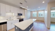 This single-story unit offers a thoughtfully designed open-concept layout that seamlessly connects the kitchen and living area. The kitchen is a standout feature, showcasing white shaker-style cabinetry extending to the ceiling, quartz countertops, and a full suite of stainless steel appliances, including a French door refrigerator, range, microwave, and dishwasher. A central island with a contrasting blue base provides additional prep space and casual seating. Recessed lighting and a linear pendant fixture illuminate the kitchen, while the adjoining living area benefits from natural light streaming through large windows and is complemented by a ceiling fan overhead. Light wood-style flooring runs throughout, lending warmth and continuity to the space. The bedroom is a calm, neutral retreat with a ceiling fan, a window allowing natural light, and ample room for furnishings. The bathroom is efficiently arranged, featuring a glass-enclosed shower, toilet, and a stacked washer and dryer tucked neatly alongside a well-organized walk-in closet with built-in shelving. The overall design is clean, modern, and functional.