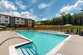 Located at 7001 Clementine Trl in Fairburn, Georgia, this outdoor amenity space is designed to deliver a resort-style experience for its residents. At the heart of the area is a generously sized swimming pool with crystal-clear water, surrounded by an expansive concrete sun deck lined with lounge chairs and market umbrellas — ideal for relaxing under the Georgia sun.

The community's clubhouse pavilion anchors one end of the pool area, featuring a striking craftsman-inspired design with exposed wood beam accents, a gabled roofline, and large windows that open the interior to natural light. Its covered porch provides a shaded retreat overlooking the pool and grounds.

On the opposite end of the pool, a dedicated outdoor grilling and dining area is enclosed by black metal fencing, outfitted with picnic-style tables and striped umbrellas, offering a convenient space for outdoor cooking and gathering. Manicured landscaping with fresh mulch beds and young plantings lines the walkways throughout the amenity area.

The property is framed by multi-story residential buildings with neutral taupe and gray exteriors, and bordered by a natural tree line that provides a lush, private backdrop. The overall atmosphere is bright, open, and well-maintained, offering a welcoming outdoor environment suited for both relaxation and social activity.