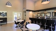 This single-floor space presents a sleek, modern interior defined by a bold black-and-white design aesthetic. The main gathering area is anchored by dramatically high ceilings that amplify the sense of openness and volume throughout the room. A distinctive horizontal black accent band runs along the upper portion of the neutral-toned walls, adding a sharp architectural detail that ties the room's color palette together.

The space is furnished with round white pedestal tables paired with classic tulip-style chairs in black, creating a retro-modern seating arrangement ideal for socializing. Statement pendant lighting with a black-and-white checkered pattern hangs from above, serving as eye-catching focal points. Large French doors along one wall allow natural light to filter in while offering a view to the exterior outdoors.

A built-in kitchen bar area lines one side of the room, featuring dark countertops, a stainless steel refrigerator, and a pair of framed mirrors mounted above. High-back black bar stools line the counter, providing additional seating. Light-toned tile flooring runs throughout, complementing the overall contrast-driven design. The combination of high ceilings, natural light, and cohesive styling gives this space a polished, inviting atmosphere well-suited for gatherings and events.