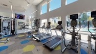 Located at 100 Heritage Pointe Rd in Charlotte, NC, this well-appointed fitness center delivers a comprehensive workout environment designed for both performance and comfort. The space is defined by soaring ceilings and expansive floor-to-ceiling windows that flood the interior with natural light while offering views of the outdoor pool and surrounding grounds. A distinctive hexagonal-patterned floor in shades of gray, blue, and yellow adds a modern, energetic aesthetic throughout.

The cardio zone features multiple treadmills and elliptical machines, each equipped with individual screens and positioned to take full advantage of the bright, window-lined wall. Ceiling fans overhead help maintain a comfortable environment during intense workouts. A large wall-mounted television provides additional entertainment for cardio sessions.

The strength and functional training area is equally well-equipped, housing a multi-functional cable machine, adjustable weight benches, a full dumbbell rack, and a colorful set of kettlebells and medicine balls neatly organized on dedicated racks. Full-length mirrors amplify the sense of space and assist with form. Motivational fitness posters and instructional displays are mounted on the walls, contributing to an informative and goal-oriented atmosphere. Glass walls and doors connect the interior to the outdoor amenity area, enhancing the sense of openness throughout.