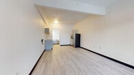 This single-floor studio unit offers a straightforward, functional layout designed to maximize its compact footprint. The main living area is finished with light-toned hardwood-style flooring throughout, complemented by clean white walls and dark baseboards that provide a crisp contrast. A structural beam detail along the ceiling adds modest architectural character to the otherwise streamlined space.

The kitchenette area is positioned along one wall and includes gray shaker-style upper and lower cabinetry, a countertop microwave, and a stainless steel refrigerator, offering essential food storage and preparation capabilities. A separate sink area with additional cabinetry is accessible through a connected alcove, providing added utility. A wall-mounted HVAC unit near the entry door ensures climate control throughout the space.

Natural light enters through a window positioned beside the front entry door, keeping the main living area bright. The bathroom is finished with light ceramic tile flooring and includes a full-size tub and shower combination, a toilet, and a vanity, all presented in a clean, neutral palette. Overall, the unit maintains a practical and well-maintained aesthetic suited for efficient single-occupancy living.