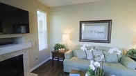 This single-story space features a thoughtfully designed living area that blends comfort with understated elegance. The room is anchored by rich, dark hardwood flooring that contrasts beautifully with the soft sage-green and warm beige wall tones, creating a calming, neutral palette throughout. A large window fitted with white horizontal blinds allows abundant natural light to filter into the space, enhancing its open and airy feel.

The living area is furnished with a plush, light seafoam-green sofa accented with decorative pillows in complementary tones, lending a relaxed yet polished aesthetic. A dark wood side table sits adjacent to the sofa, topped with a classic white table lamp and a small potted green plant, adding a touch of warmth and life to the setting. A large framed artwork with a dark espresso-toned border hangs prominently on the wall, serving as a refined focal point.

White orchids in the foreground introduce an elegant floral element, reinforcing the space's serene and welcoming ambiance. Clean white baseboards and smooth walls complete the well-maintained interior, making this a comfortable and visually cohesive living environment.