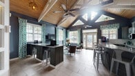 This single-story community space makes a striking architectural impression with its dramatic vaulted cathedral ceiling, where exposed white-painted structural beams contrast beautifully against warm natural wood planking. Ceiling fans suspended from the peaks provide airflow throughout the open layout, while clerestory windows near the roofline flood the interior with abundant natural light.

The main gathering area features large windows dressed in teal patterned curtains, offering outdoor views and reinforcing the coastal-inspired color palette of deep navy walls, grey tones, and aqua accents. A comfortable lounge seating arrangement—anchored by a geometric-patterned area rug—includes upholstered chairs positioned around a central coffee table, with wall-mounted televisions nearby. A reception or bar-height counter with dark granite tops and industrial-style metal stools provides additional functional space.

An adjoining kitchen area is well-appointed with grey cabinetry, granite countertops, a white subway tile backsplash, stainless steel appliances, and a Lavazza coffee station. French doors throughout connect the interior to the outdoors, enhancing the sense of openness. Light-toned large-format tile flooring runs throughout, completing the cohesive, inviting atmosphere of this versatile amenity space.