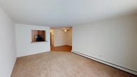 This single-floor residence offers a practical and straightforward layout designed for comfortable everyday living. The main living area is finished with wall-to-wall carpeting in a neutral beige tone, providing a warm and understated foundation for the space. Cream-colored walls throughout contribute to a clean, bright atmosphere that feels open and easy to personalize.

A defining architectural feature is the pass-through window connecting the living area to the kitchen, offering both visual openness and functional convenience. The kitchen, visible through the pass-through, is equipped with dark wood cabinetry and stainless steel appliances, including a refrigerator and microwave, giving it a practical and polished appearance.

Adjacent to the carpeted living space, a secondary area features hardwood-style flooring and is equipped with a ceiling fan, providing ventilation and adding a subtle design element overhead. A baseboard heating unit runs along the exterior wall, ensuring efficient climate control throughout the cooler months. A hallway visible in the background suggests additional rooms beyond the main living space, hinting at a well-organized and functional floor plan overall.