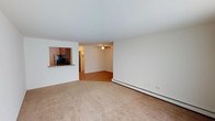 This single-floor residence offers a practical and straightforward layout designed for comfortable everyday living. The main living area is finished with wall-to-wall carpeting in a neutral beige tone, providing a warm and understated foundation for the space. Cream-colored walls throughout contribute to a clean, bright atmosphere that feels open and easy to personalize.

A defining architectural feature is the pass-through window connecting the living area to the kitchen, offering both visual openness and functional convenience. The kitchen, visible through the pass-through, is equipped with dark wood cabinetry and stainless steel appliances, including a refrigerator and microwave, giving it a practical and polished appearance.

Adjacent to the carpeted living space, a secondary area features hardwood-style flooring and is equipped with a ceiling fan, providing ventilation and adding a subtle design element overhead. A baseboard heating unit runs along the exterior wall, ensuring efficient climate control throughout the cooler months. A hallway visible in the background suggests additional rooms beyond the main living space, hinting at a well-organized and functional floor plan overall.