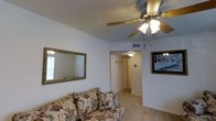This single-floor residence offers a comfortable and functional layout with a warm, lived-in character. The living area serves as the heart of the space, featuring light-toned carpeted flooring, textured ceilings, and a ceiling fan with an integrated light fixture that provides both airflow and ambient lighting. The walls are painted in a clean, neutral white that helps reflect light and contributes to an open feel. A large ornate-framed mirror adorns one wall, enhancing the sense of space and adding a classic decorative touch, while a framed artwork piece provides additional visual interest. A hallway visible from the living area connects to the rest of the home, with a ceiling-mounted light fixture guiding the way. Electrical outlets and switches are thoughtfully placed along the walls, and a ceiling vent indicates a central climate control system. The overall ambiance is cozy and understated, with a traditional aesthetic reflected in details such as the floral-patterned sofa and gold-toned accents. The layout flows naturally, offering a practical and welcoming environment suitable for everyday living.
