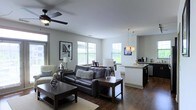 This single-floor space showcases an open-concept layout that seamlessly connects the living room, dining area, and kitchen into one cohesive, functional environment. The living area is anchored by a dark leather sofa and loveseat arrangement, complemented by a wood coffee table and area rug that define the space. A ceiling fan with light fixture overhead provides both comfort and illumination. Multiple windows along the walls flood the interior with natural light, enhancing the clean, neutral-toned walls.

Adjacent to the living area, a dining space accommodates a wooden table and chairs, positioned near additional windows that continue the bright, airy feel throughout. The kitchen features dark cabinetry, granite or stone countertops, and pendant lighting suspended above the counter, adding a refined touch. Dark hardwood flooring runs consistently throughout the space, creating visual continuity and warmth. Wall art and carefully selected décor add personality without overwhelming the overall modern, understated aesthetic. The open flow between areas makes the space feel expansive and well-suited for both everyday living and entertaining.