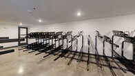 Located at 1803 S 46th St in Rogers, Arkansas, this purpose-built bike storage room offers a well-organized and functional space designed to accommodate a large number of bicycles. The room spans a generous single floor and features an L-shaped layout, as seen in the overhead dollhouse view, maximizing usable wall and floor space throughout.

The interior is lined with rows of heavy-duty, black metal vertical bike racks arranged in parallel configurations, providing efficient and secure storage capacity for numerous bicycles. The racks are sturdy in construction and designed to hold bikes at varying heights, making efficient use of the room's vertical space.

The flooring consists of polished concrete, offering a durable, low-maintenance surface well-suited for high-traffic utility use. The walls are finished in clean white paint, contributing to a bright and open atmosphere. Recessed ceiling lighting provides consistent, even illumination throughout the room, while a ceiling-mounted sprinkler system and security camera indicate attention to safety and security.

Ventilation is supported by a ceiling-mounted HVAC vent, ensuring air circulation within the enclosed space. A single access door with a glazed panel provides entry to the room. Overall, this bike storage facility is a practical, well-appointed amenity built to serve the needs of an active residential or commercial community.
